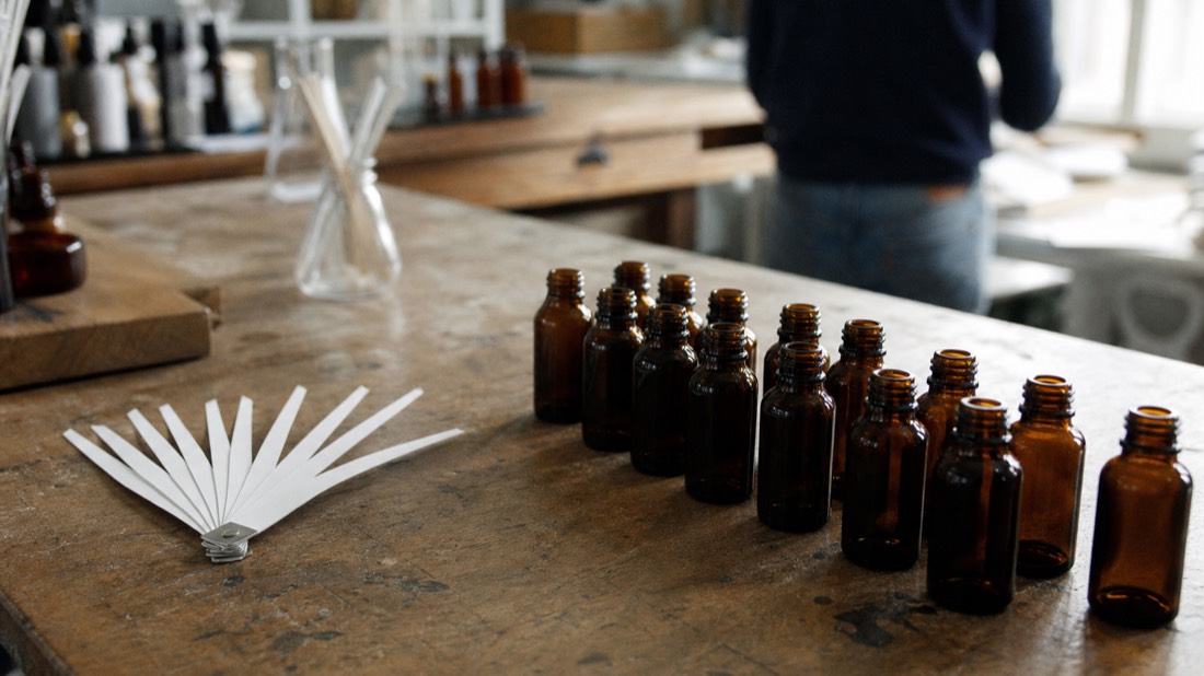 Fan of blank paper scent strips beside rows of small amber bottles on a workshop bench in soft daylight