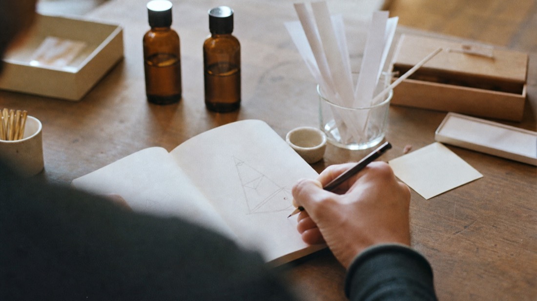 Hand sketching a fragrance pyramid in a notebook beside scent strips at a perfume workshop table