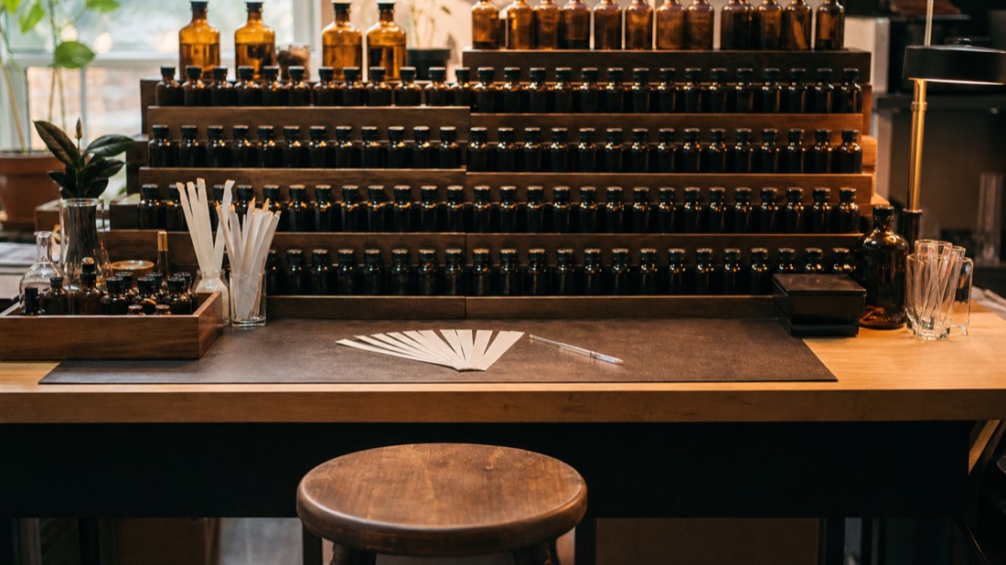 Empty wooden stool facing a stocked tiered rack of essence bottles with fresh paper scent strips laid out