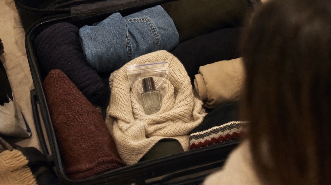 Perfume bottle wrapped in a soft sweater and sealed in a zip bag, nested in the middle of a packed suitcase