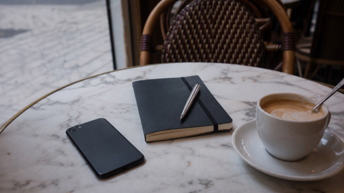 Phone lying on a Paris cafe table beside a small notebook and coffee, checking a workshop listing before booking