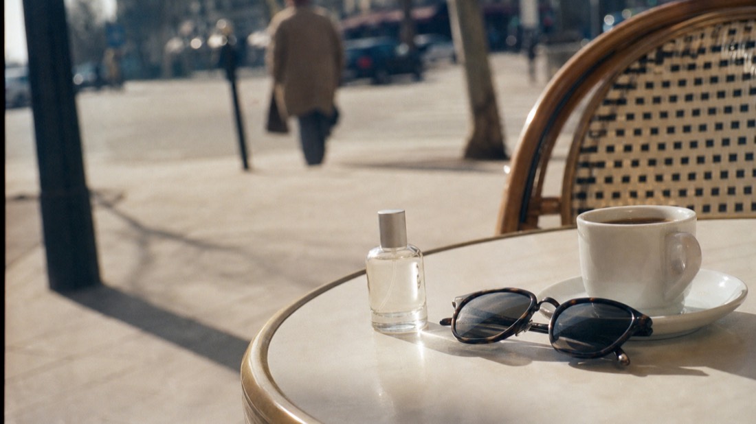 Small unbranded perfume bottle and sunglasses on a cafe table with a blurred Paris avenue behind, planning a workshop day