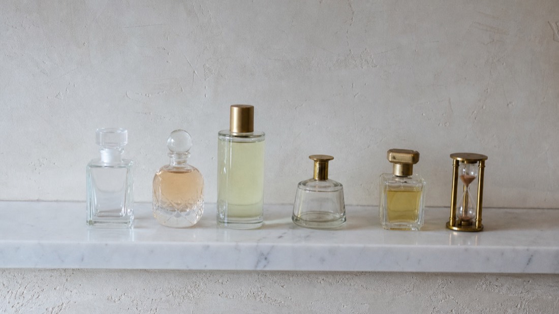 Five unlabeled perfume flacons of different sizes beside a small hourglass on a marble shelf, comparing Paris workshop durations