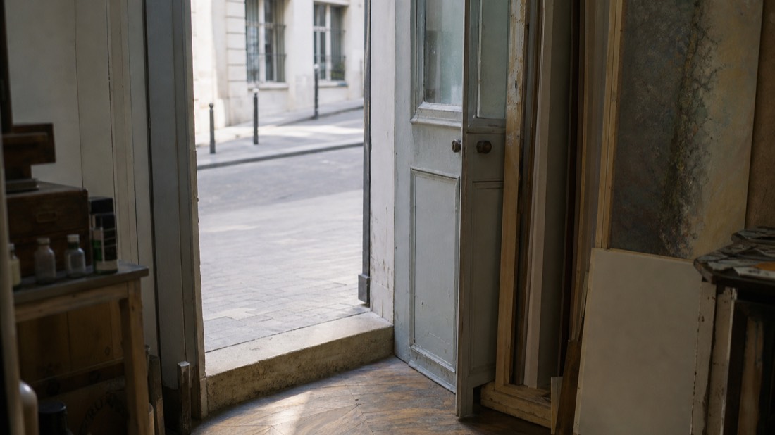 Half-open door of a small Paris atelier with a step at the threshold and daylight falling across the workshop floor